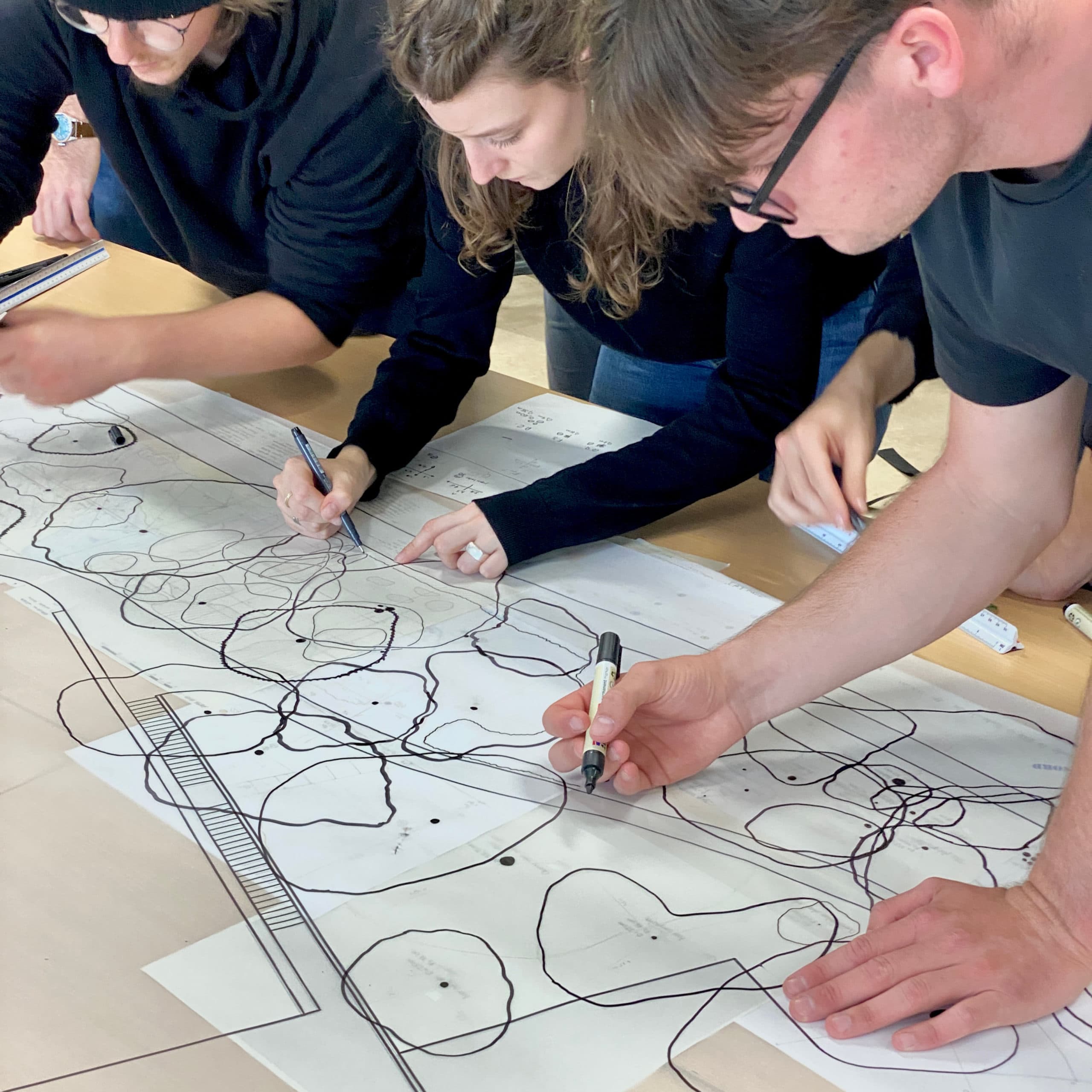 Gros plan sur plusieurs participants travaillant en équipe autour d'une table, traçant au feutre des annotations et des formes sur un grand plan imprimé lors d'un atelier collaboratif.